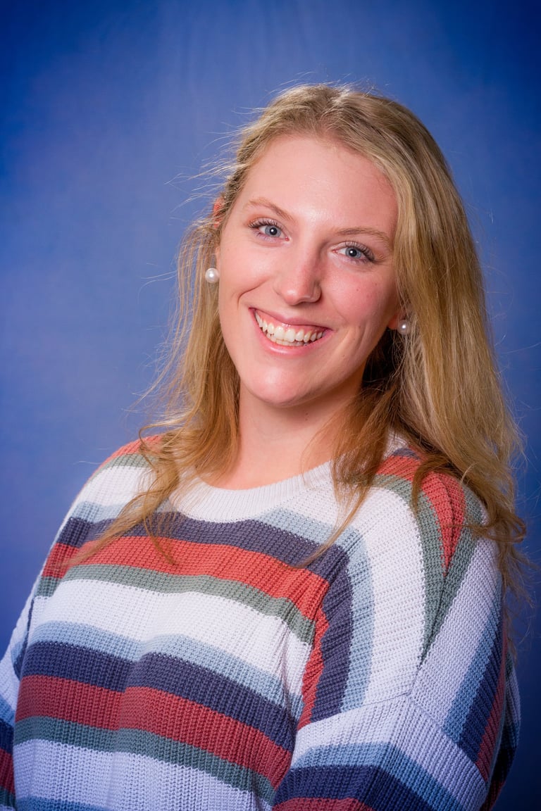 Young woman with long blonde hair smiling at camera, wearing a red, white, and blue striped sweater against a blue background